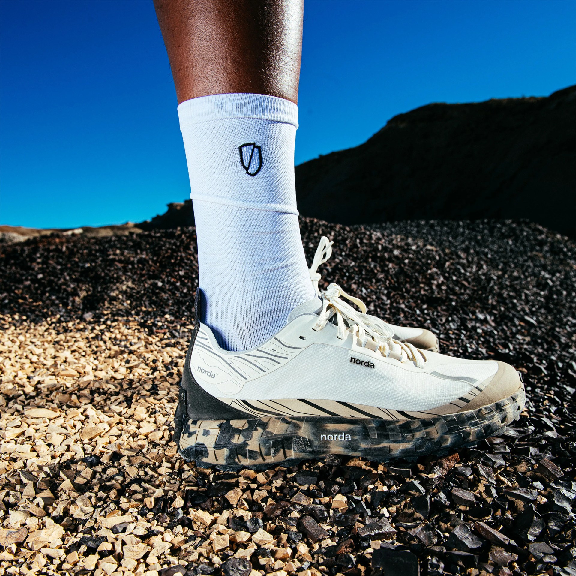 White and beige Norda trail running shoe with black patterned sole worn with white socks on rocky ground.
