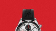 Silver chronograph watch with black bezel and subdials, black textured rubber strap, on red background.