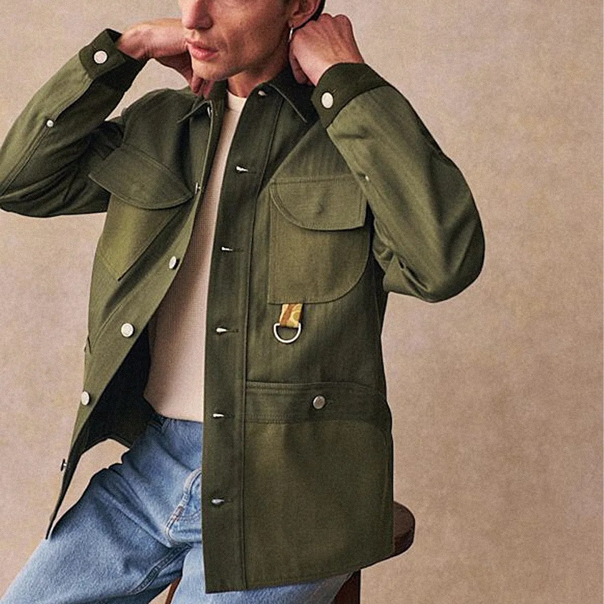 Olive green button-up jacket with flap pockets and a small yellow strap detail worn over a white shirt and blue jeans.
