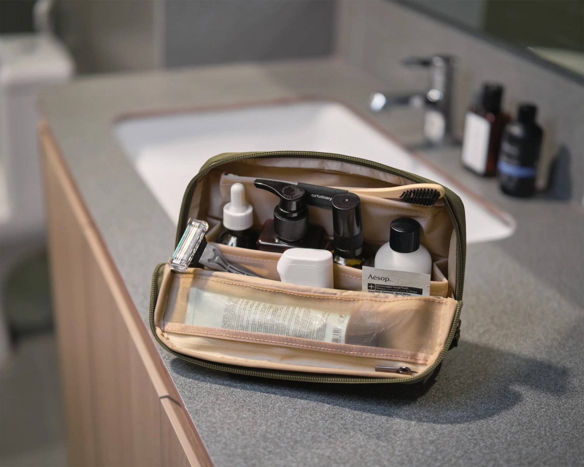 Olive green toiletry bag with beige interior holding grooming items on a gray bathroom countertop.