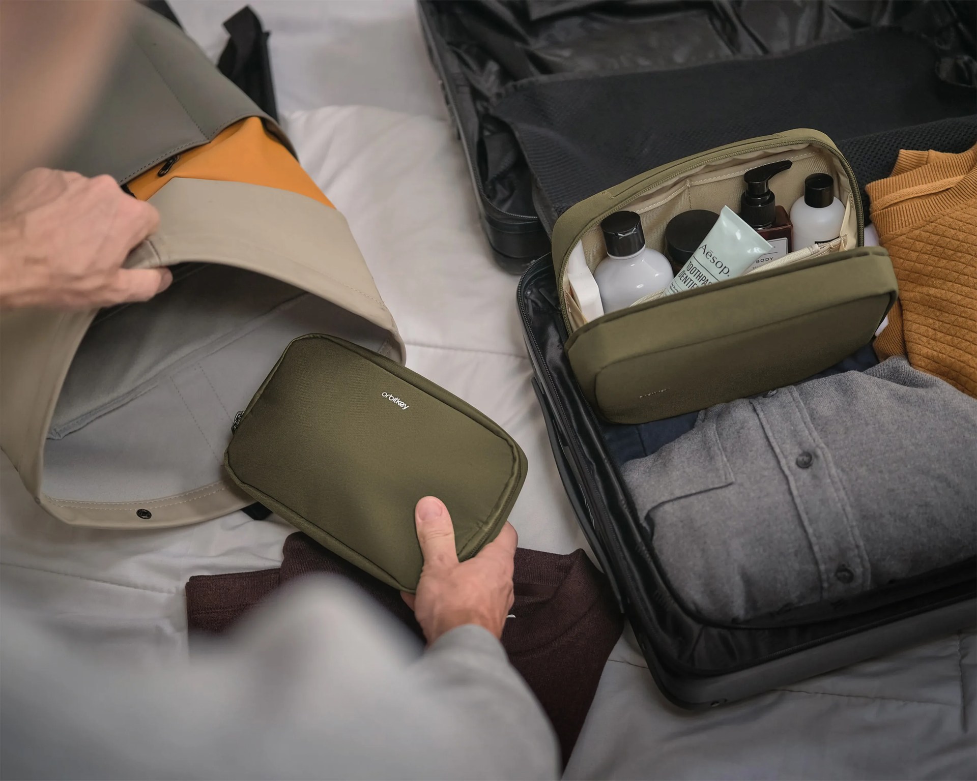 Olive-green Orbitkey toiletry bag being packed into a beige and orange backpack next to an open suitcase with clothes and toiletries.