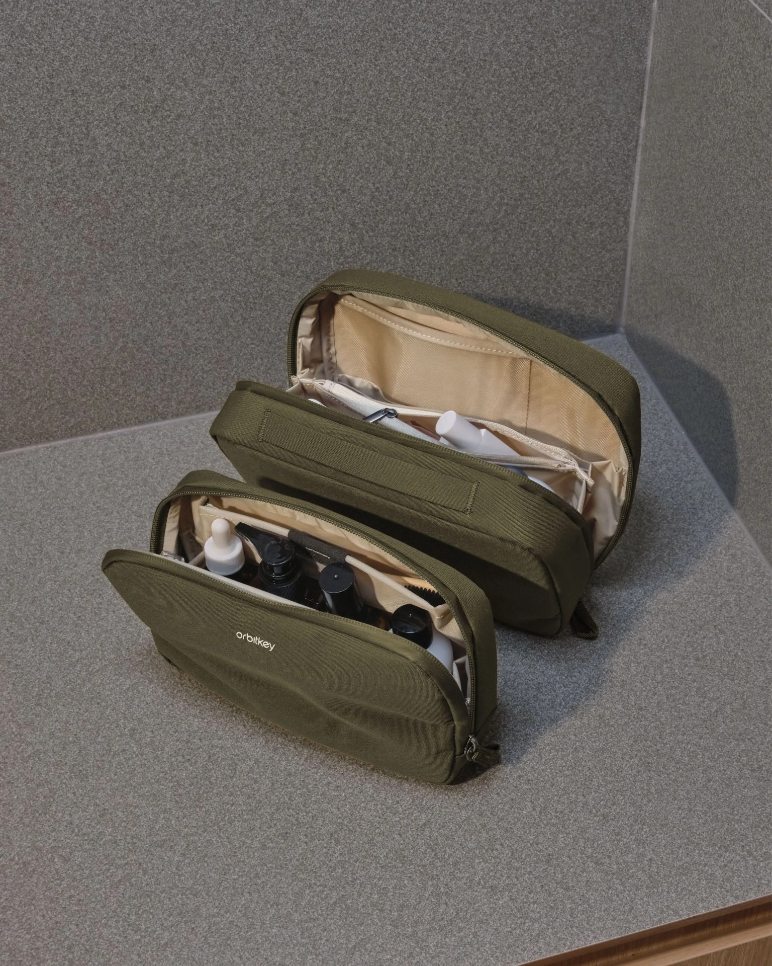 Two olive-green fabric toiletry bags with beige interiors, partially unzipped and filled with bottles, on a gray countertop.