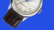 Silver watch with a cream dial, blue hands, Roman numerals, and a dark brown leather strap on a blue background.