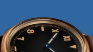 Close-up of a black watch face with beige Roman and Arabic numerals and blue hands under a domed crystal.