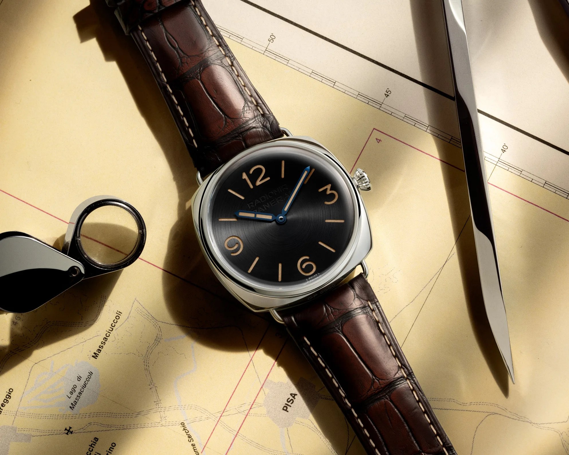 Silver wristwatch with black dial, tan numerals, blue hands, and brown leather strap on a map background.
