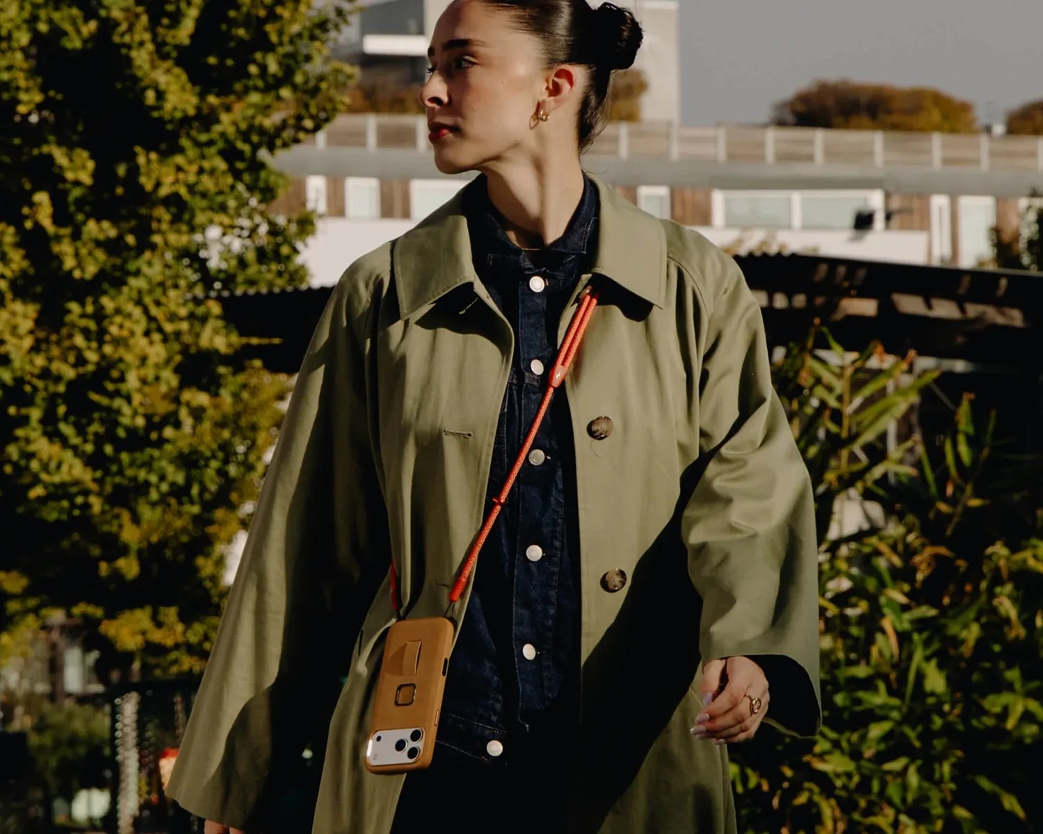 Person wearing an olive green coat over a dark denim jacket with a tan phone case hanging from an orange strap.
