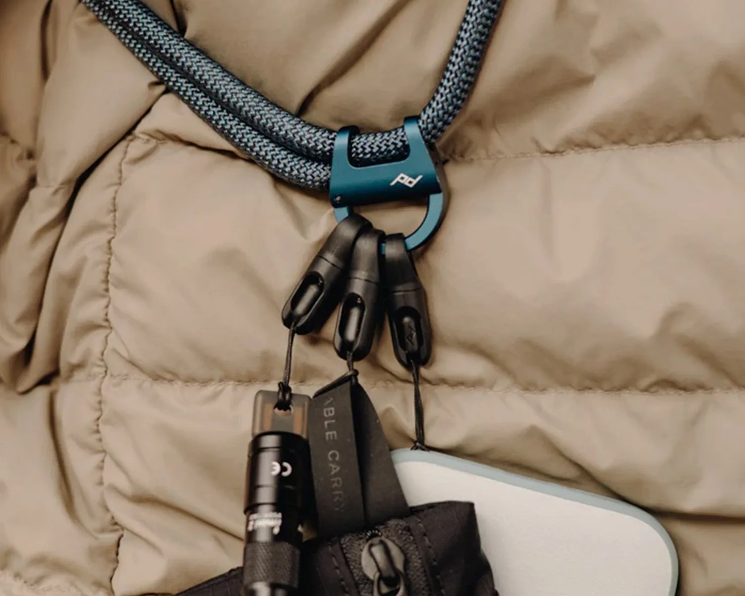 Blue metal carabiner with three black plastic clips attached to a gray and black braided cord on a beige quilted fabric.