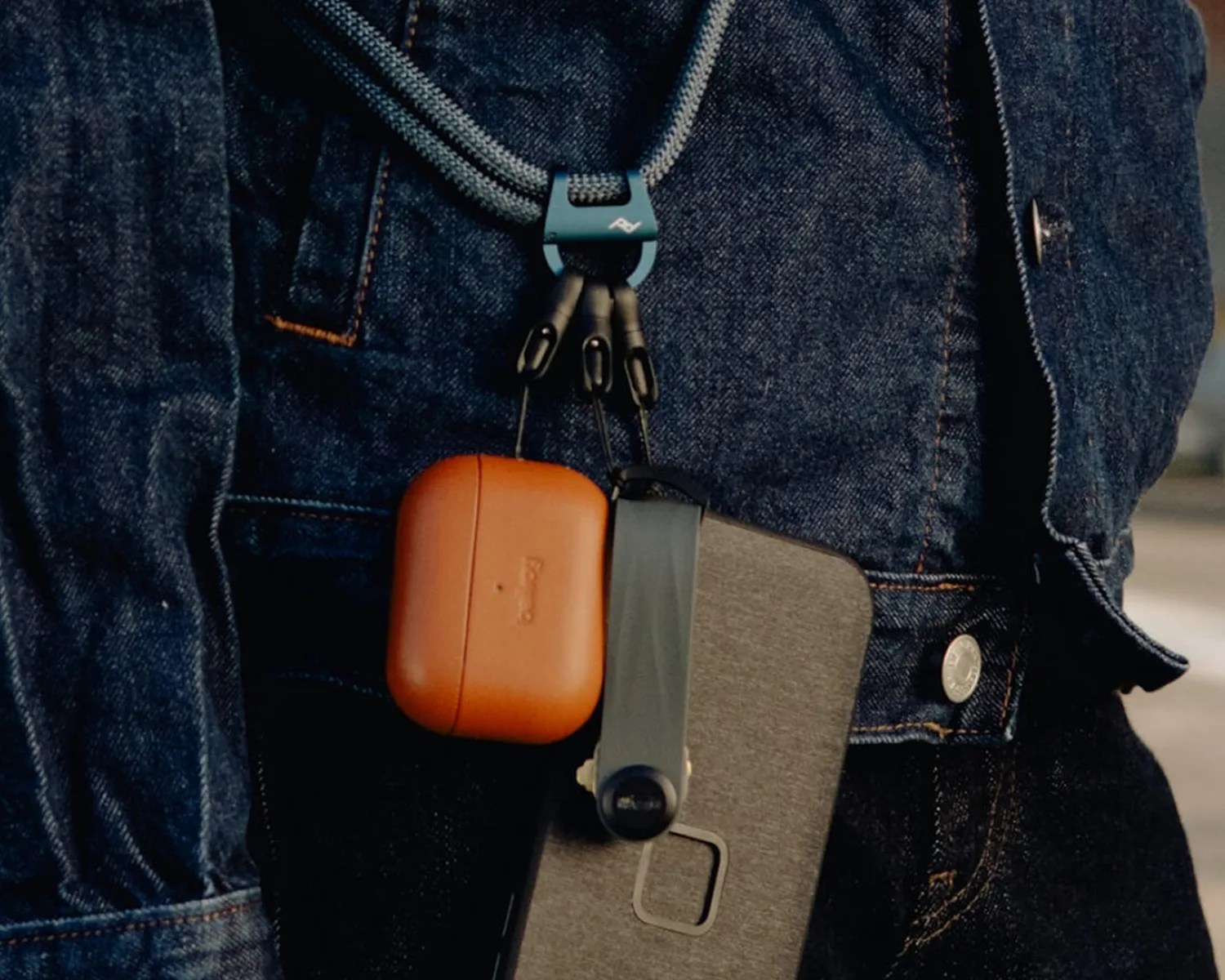 Brown leather AirPods case and gray fabric phone case attached to a blue cord worn over a denim jacket.