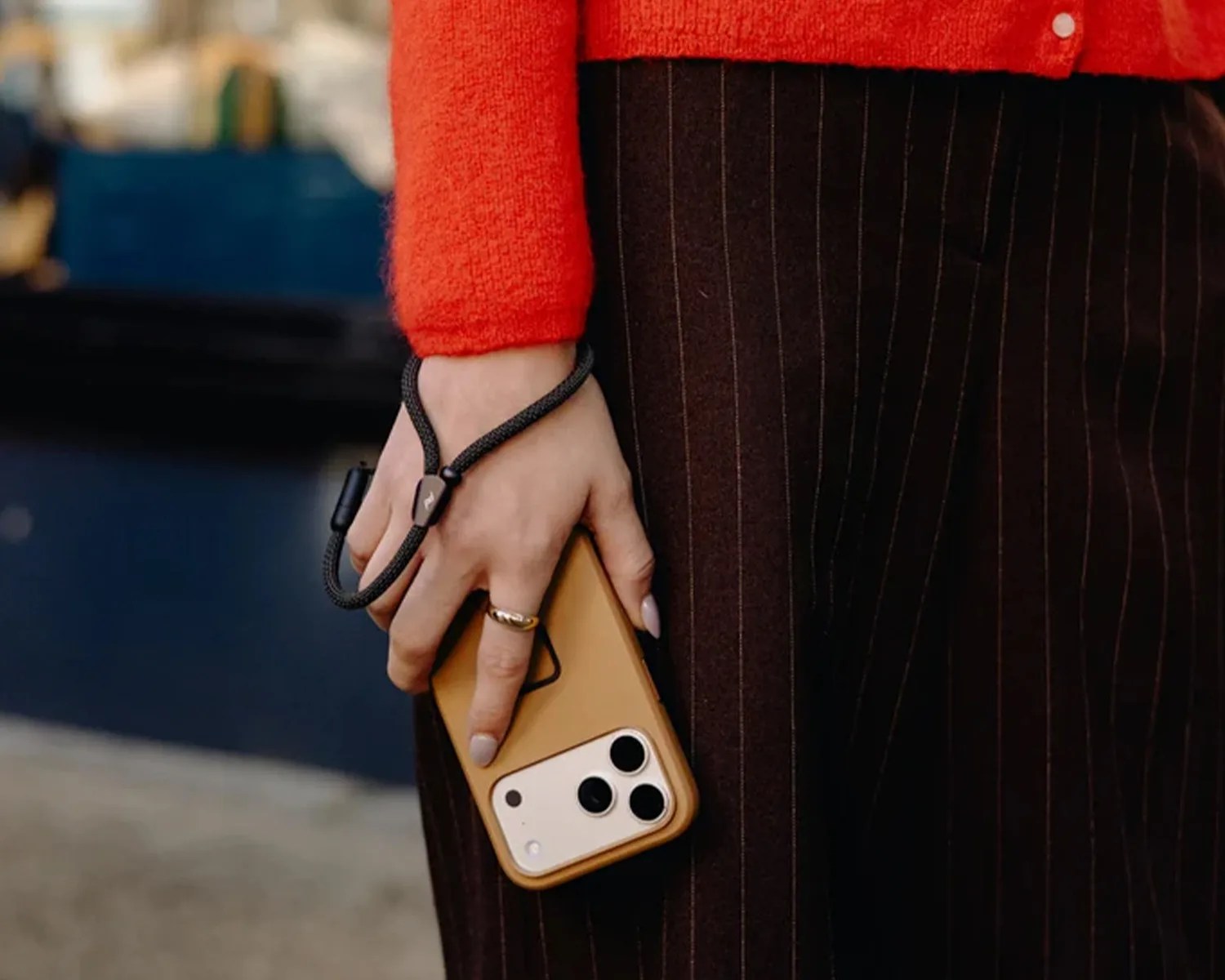 Hand holding a smartphone with a tan case and black wrist strap, wearing a gold ring and red sweater.
