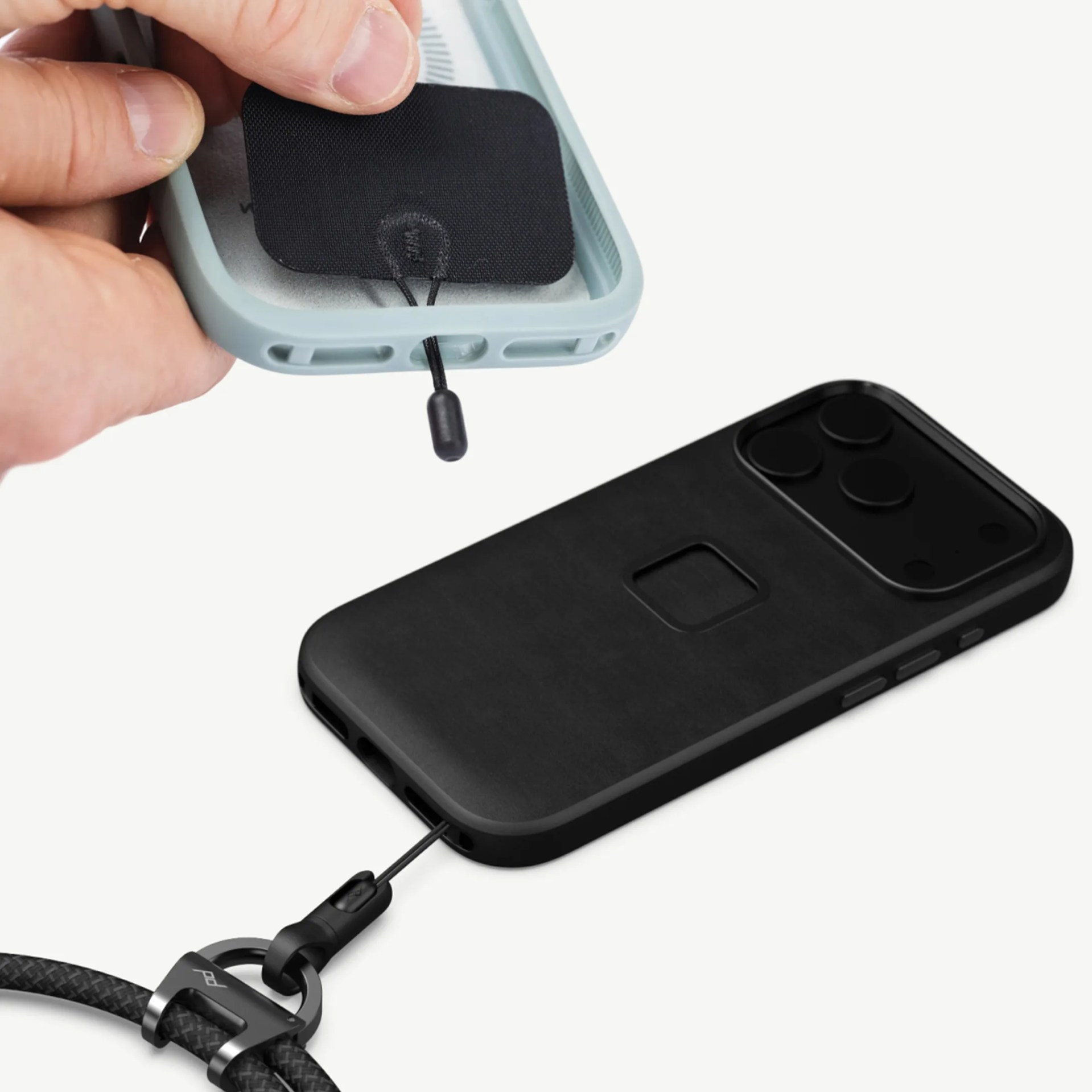 Black phone case with a camera cutout and attached black wrist strap, with a hand placing a black adhesive patch inside a light blue phone case.