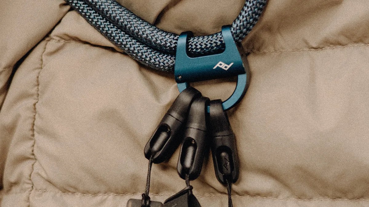 Blue metal carabiner with a white logo attached to a black and gray braided rope on a beige quilted fabric.