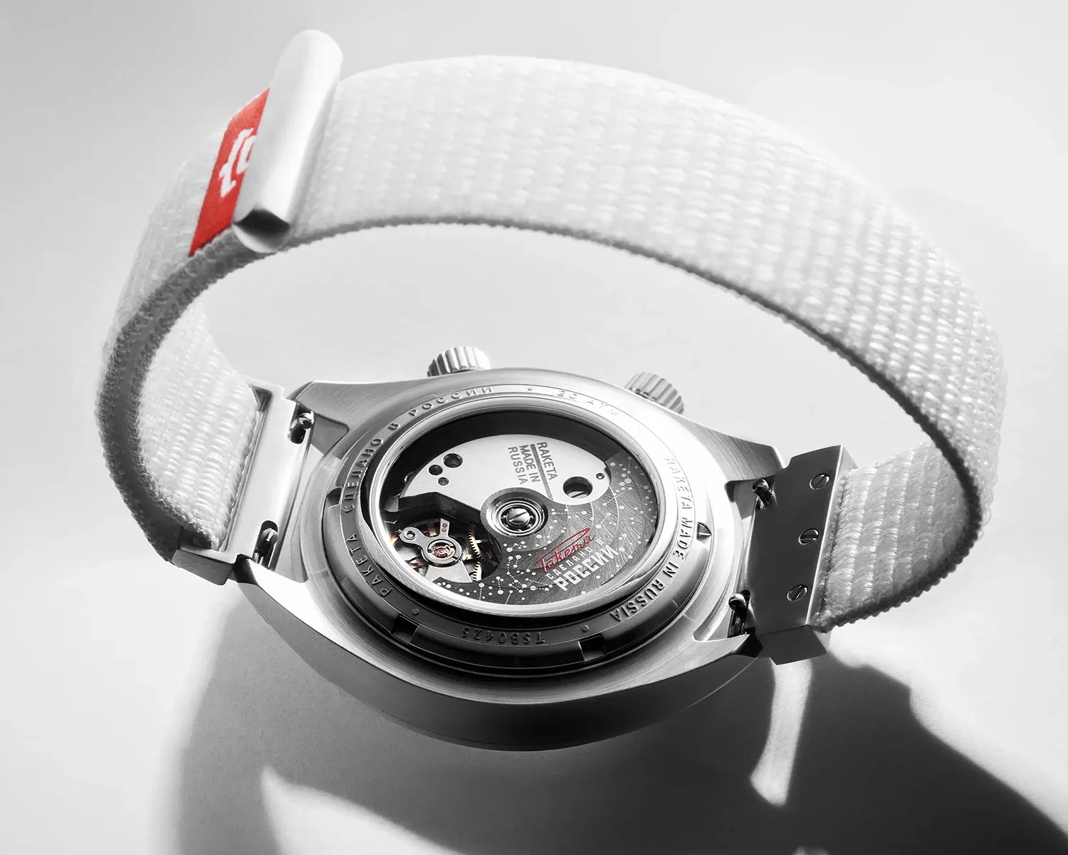 Silver Raketa watch with visible mechanical movement and white woven strap with red tag.