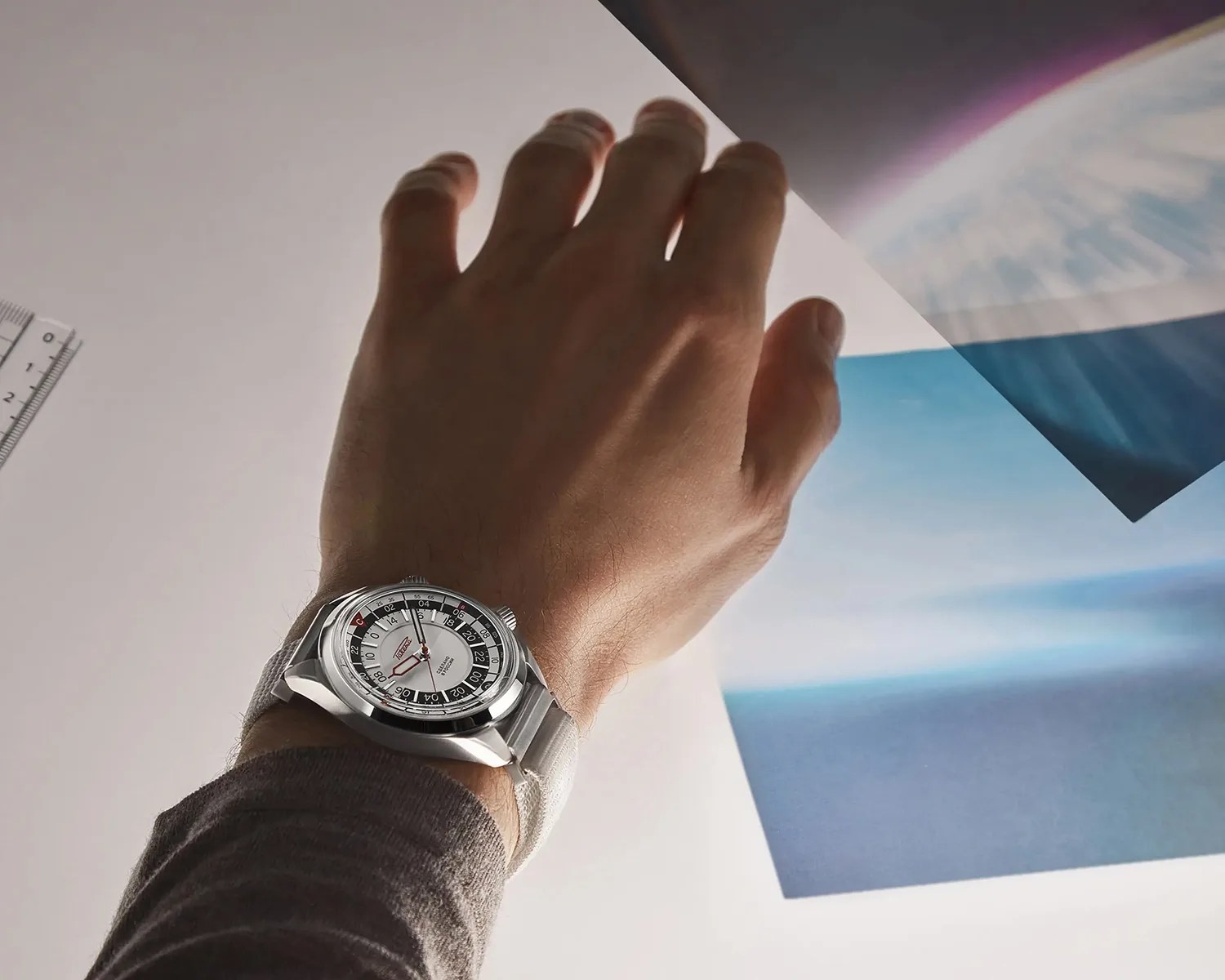 Silver wristwatch with a white dial and black and red accents worn on a wrist over a gray sleeve.