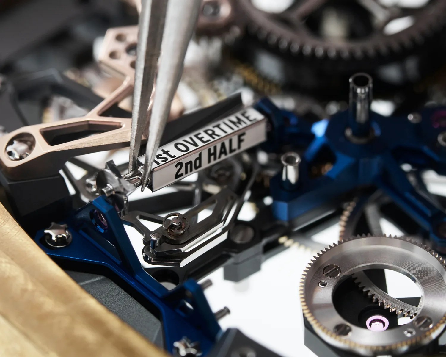 Close-up of watch mechanism with tweezers adjusting a component labeled "1st OVERTIME 2nd HALF" among blue and metallic gears.