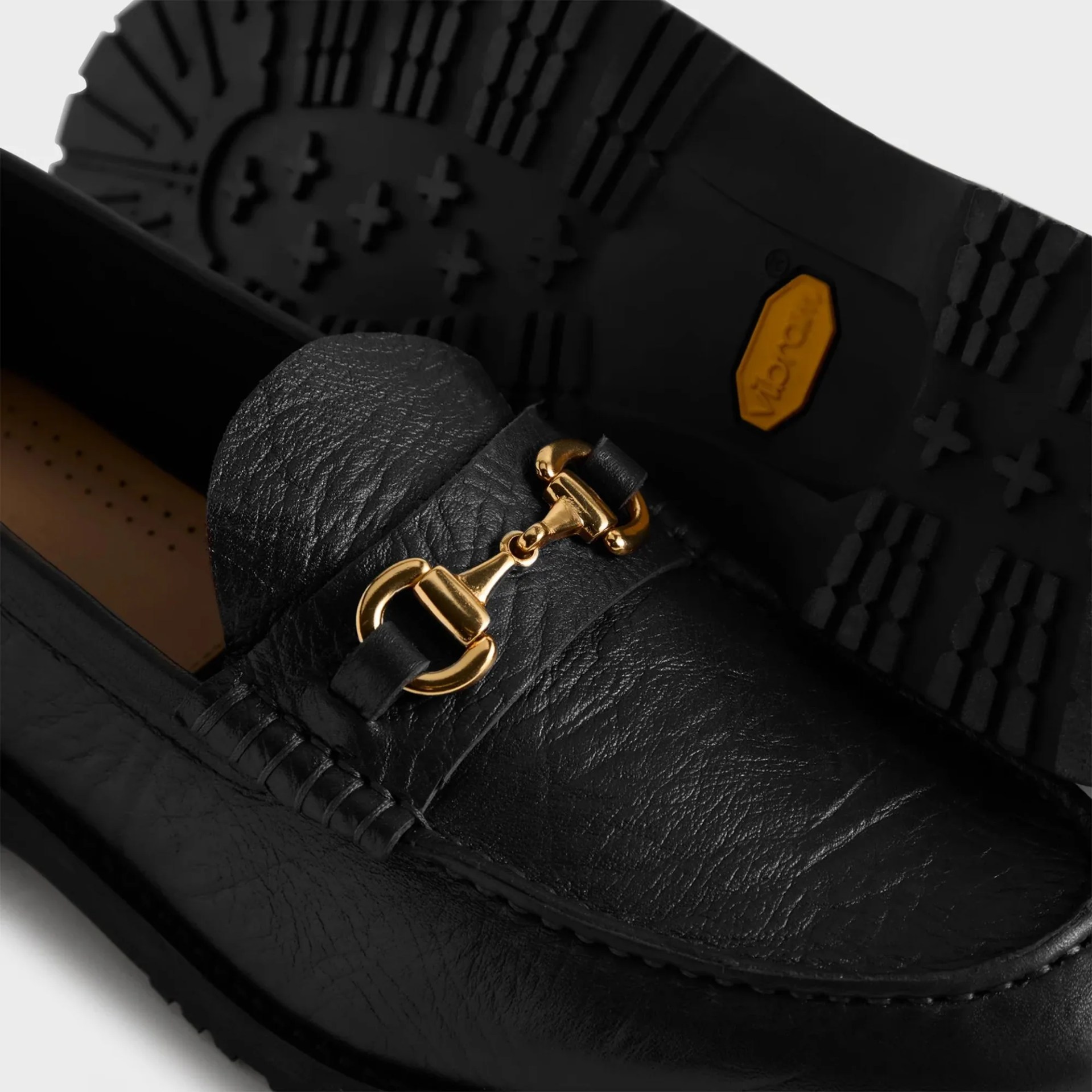 Black leather loafer with gold horsebit detail and rugged Vibram sole visible in the background.