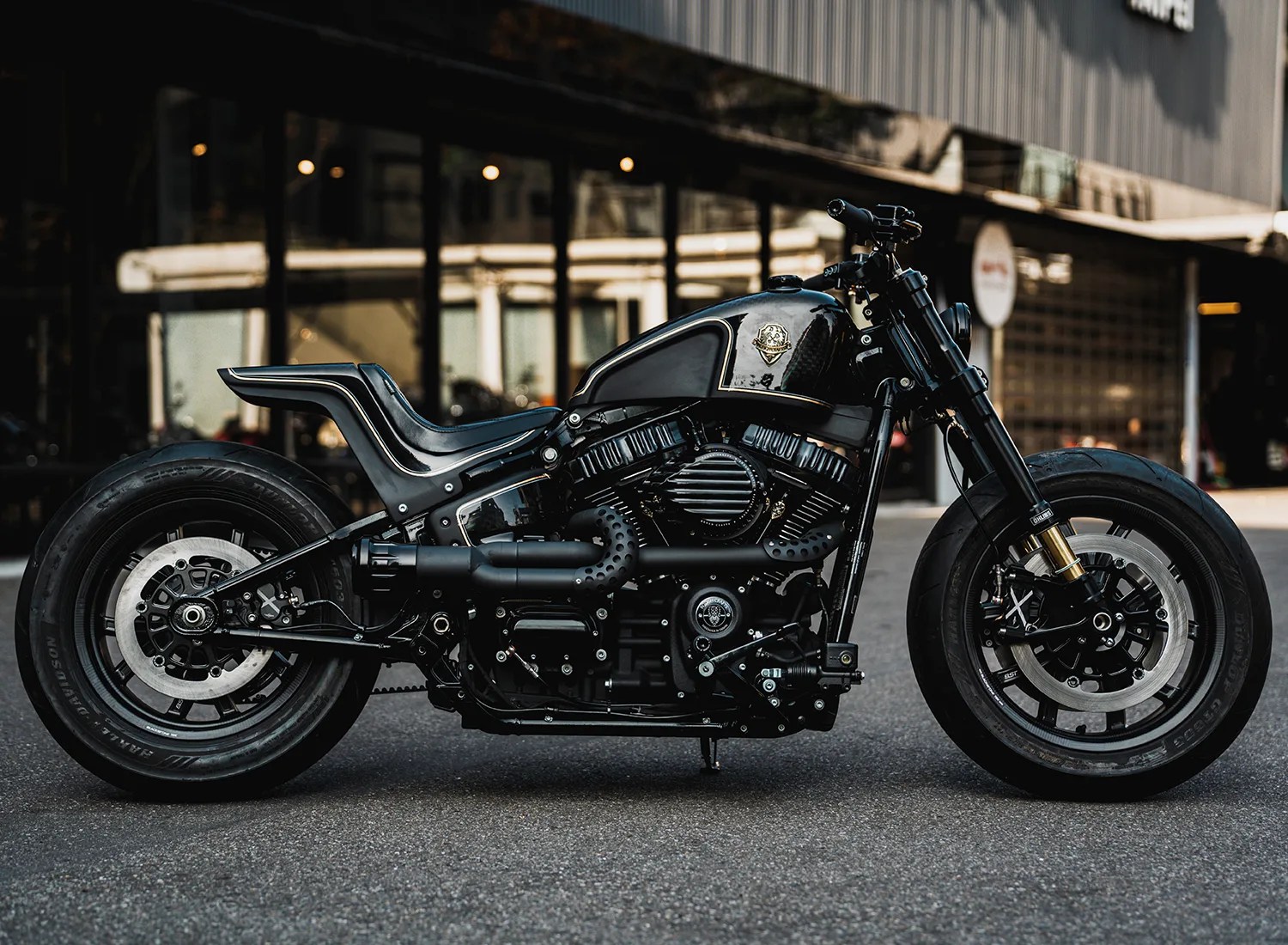 Black custom motorcycle with a matte and glossy finish parked on asphalt in front of a building with large windows.