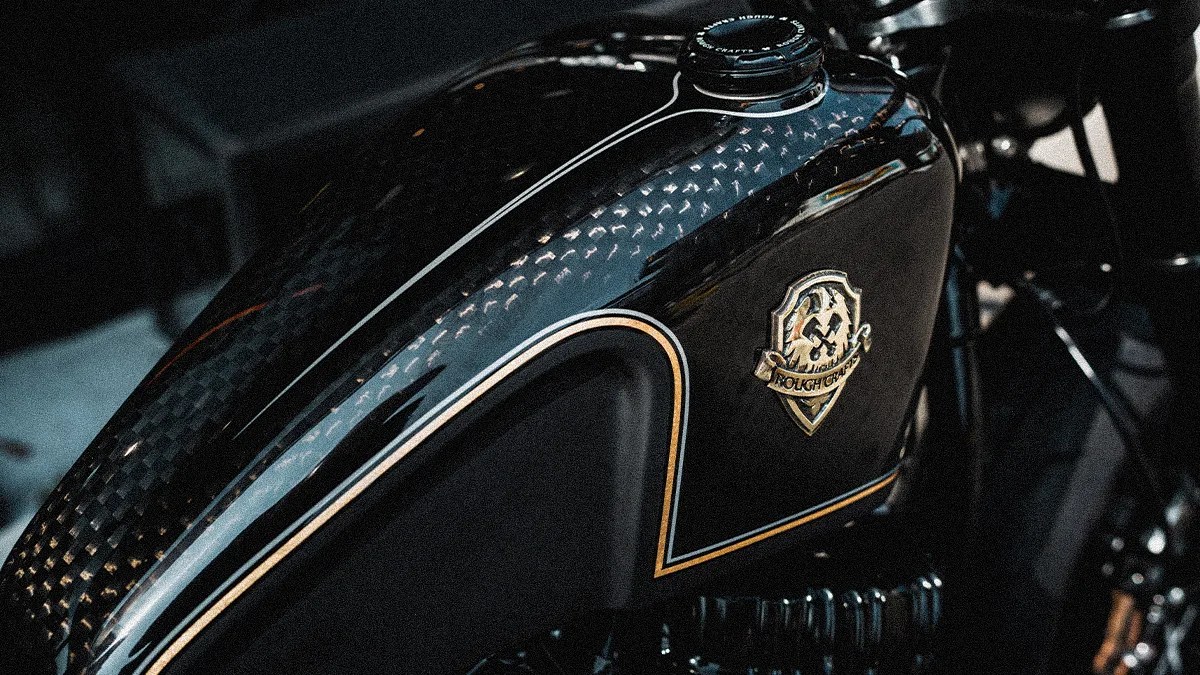 Glossy black motorcycle fuel tank with gold trim and a metallic emblem on the side.