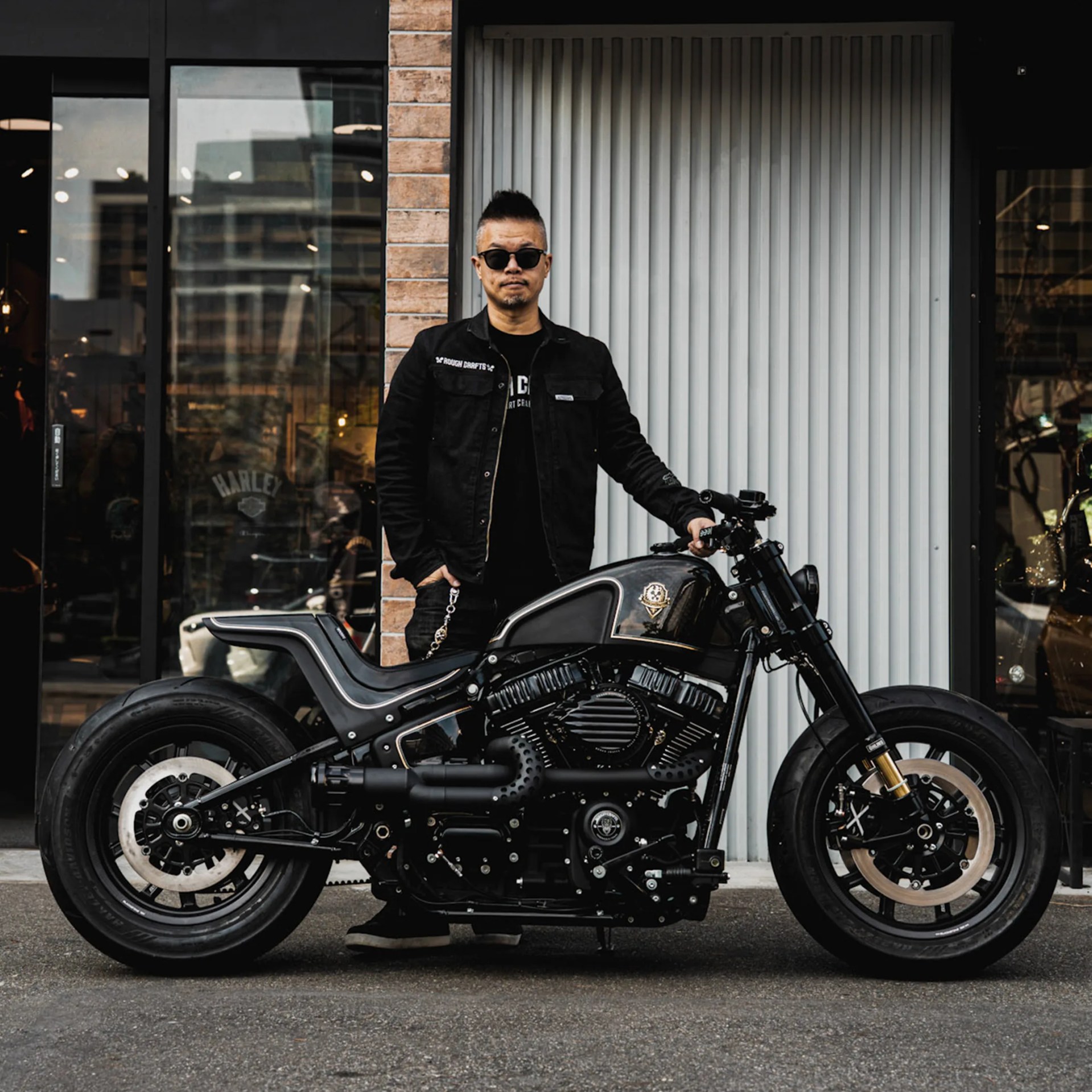 Man in black jacket and sunglasses standing next to a matte black custom motorcycle in front of a building.
