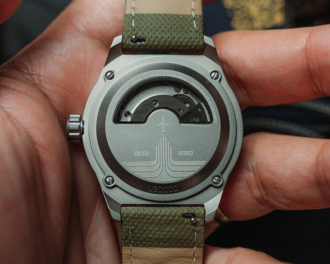 Back of a titanium wristwatch with visible automatic movement, green textured strap, and engraved airplane design.
