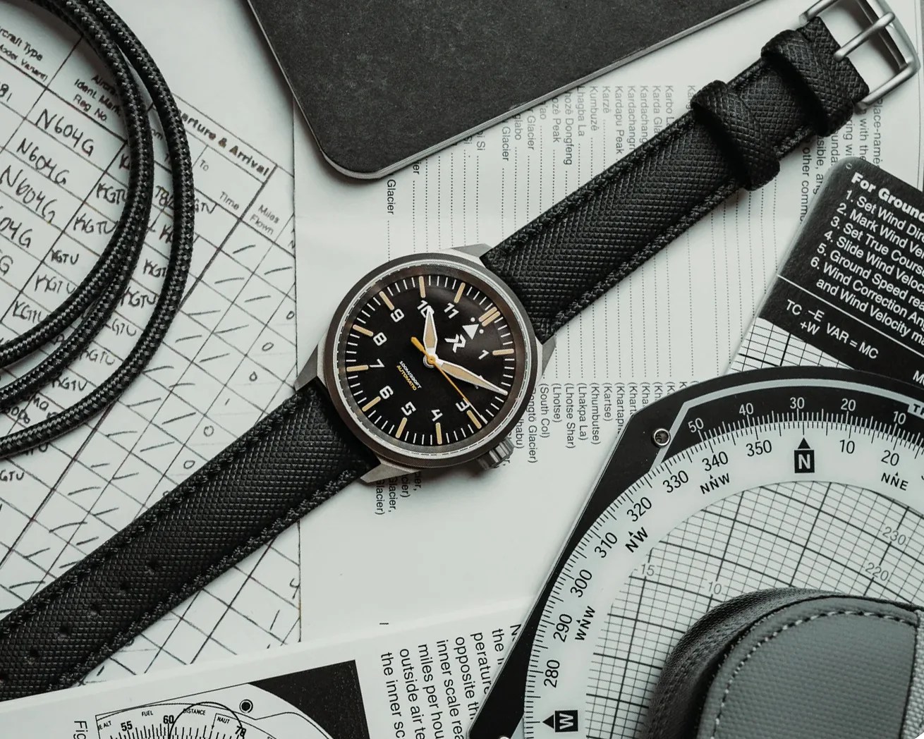Black wristwatch with a textured strap and black dial showing 3:55 on top of aviation charts and a flight computer.