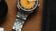 Stainless steel wristwatch with an orange textured dial and black-and-white hour markers on a brown surface.