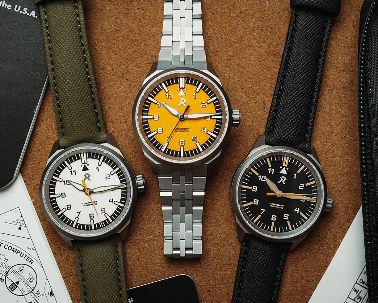 Three wristwatches with round faces: one with an orange dial and silver metal bracelet, one with a white dial and green fabric strap, and one with a black dial and black fabric strap, all on a corkboard surface.