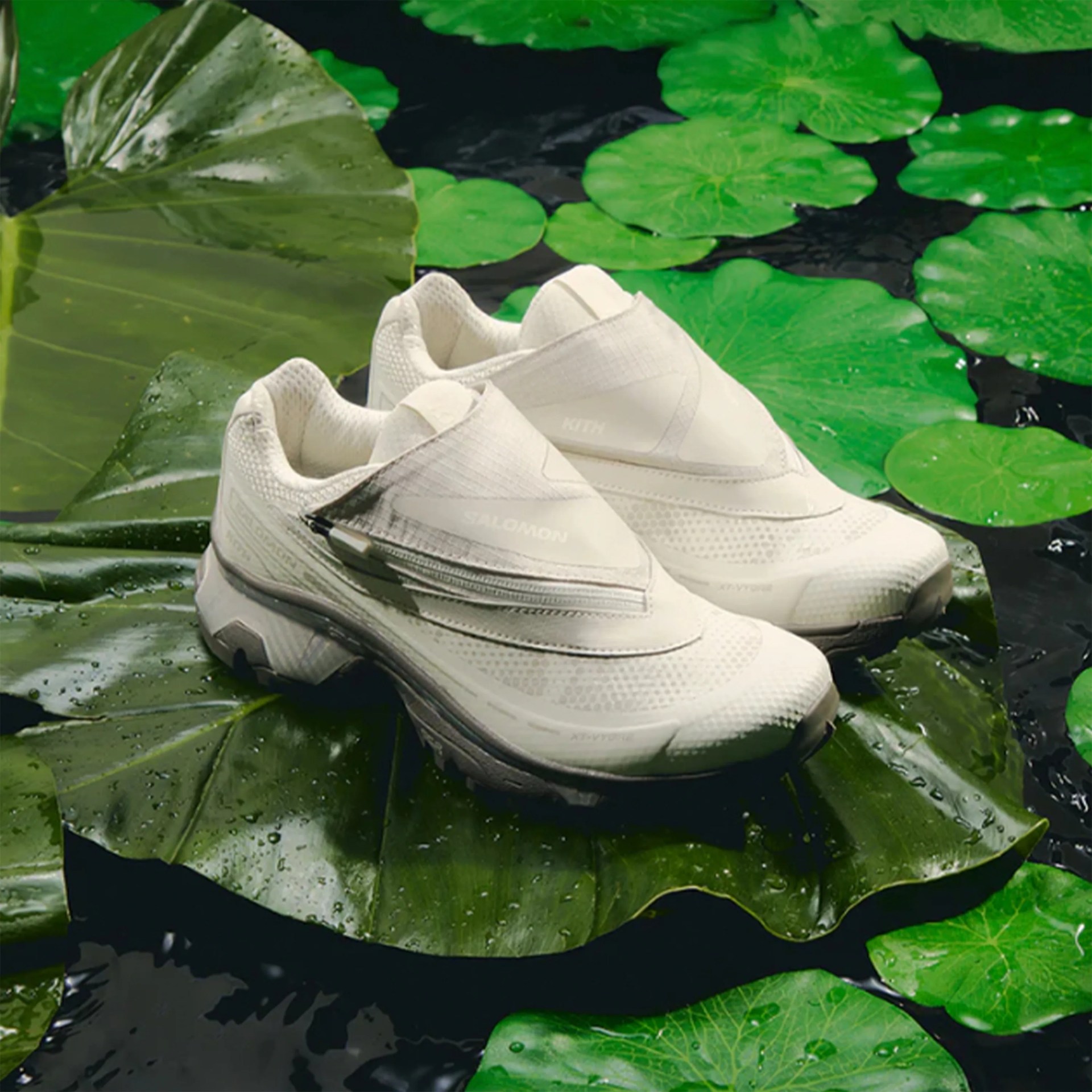 Pair of white Salomon sneakers with a zippered cover, placed on large green lily pads floating on water.