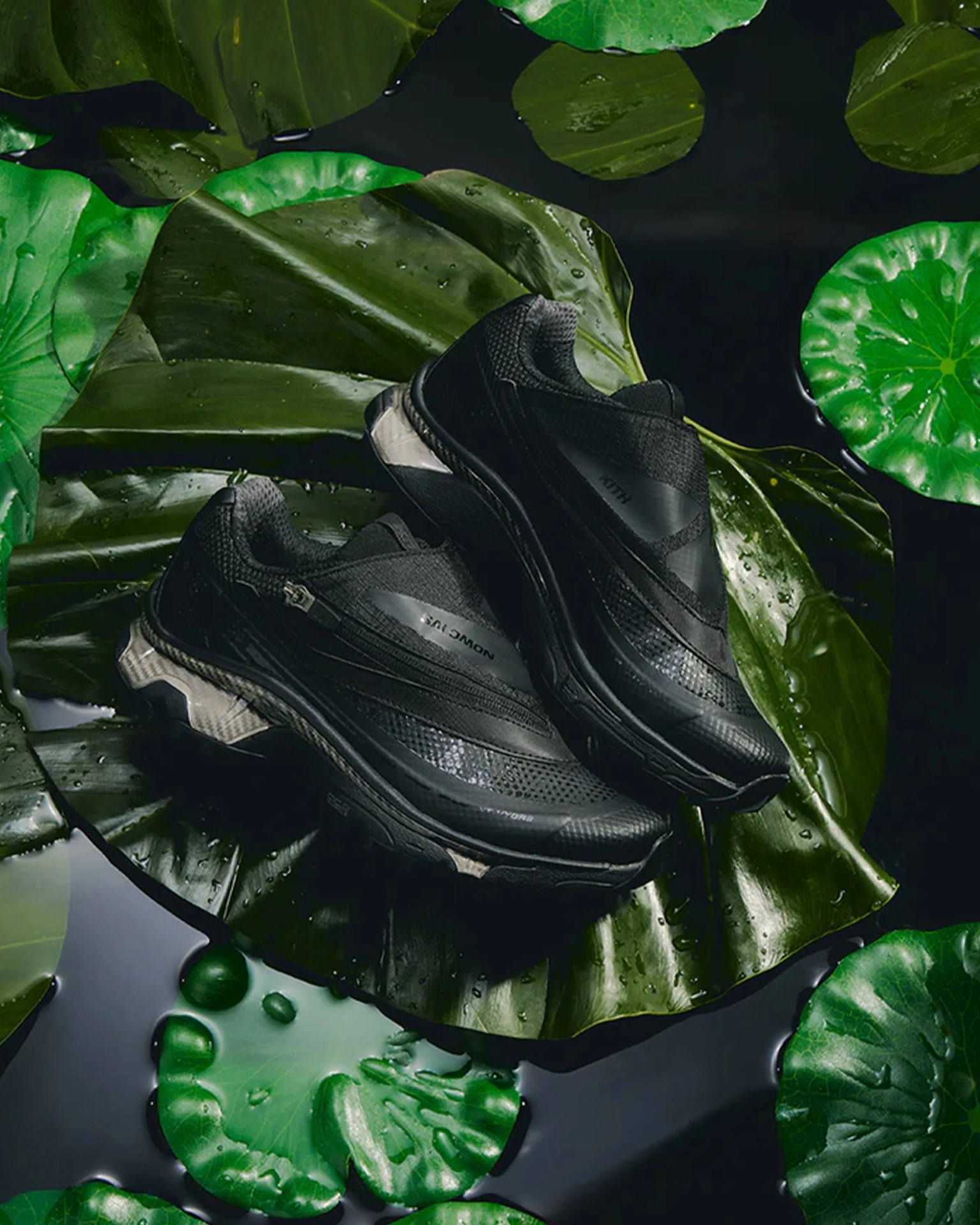 Pair of black athletic sneakers with textured and smooth panels resting on large green lily pads floating on dark water.