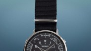 Close-up of a Seiko watch with a black dial, silver case, and black fabric strap against a blue gradient background.