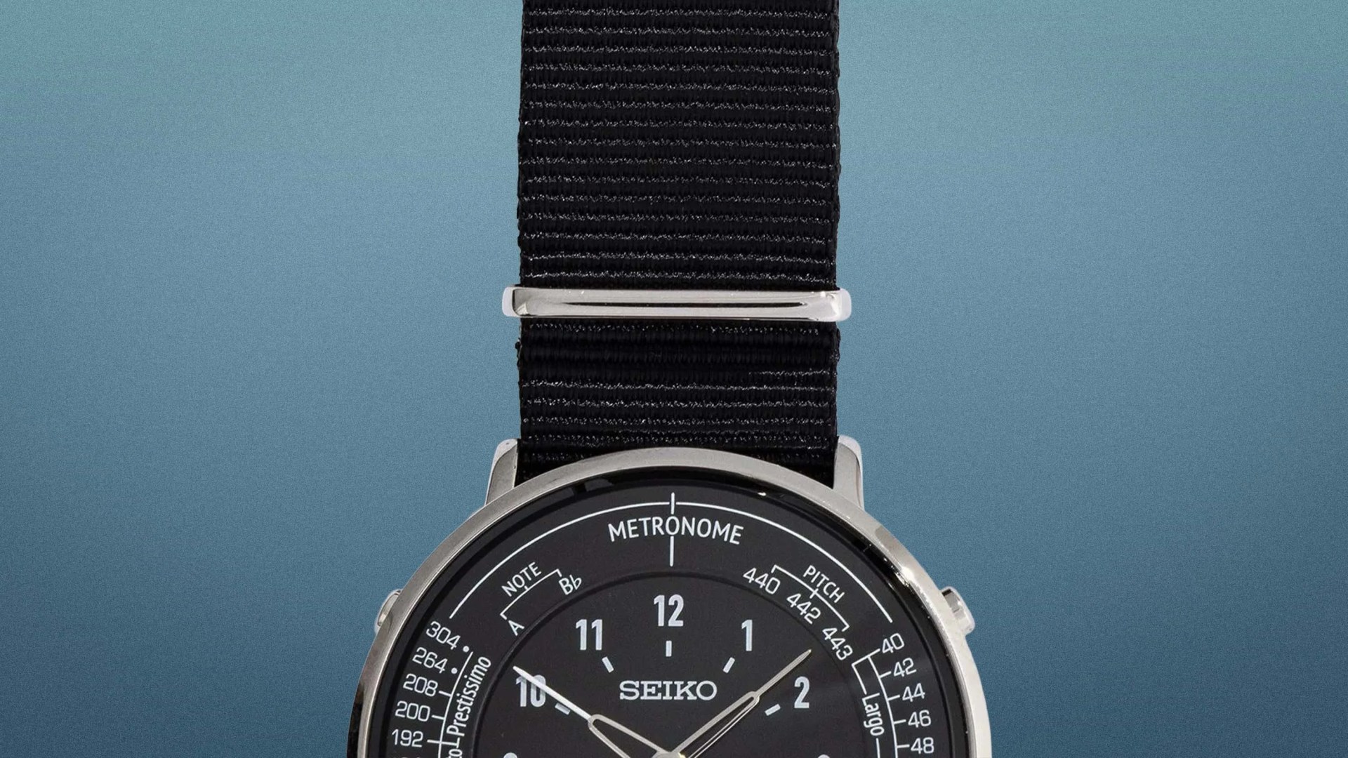 Close-up of a Seiko watch with a black dial, silver case, and black fabric strap against a blue gradient background.