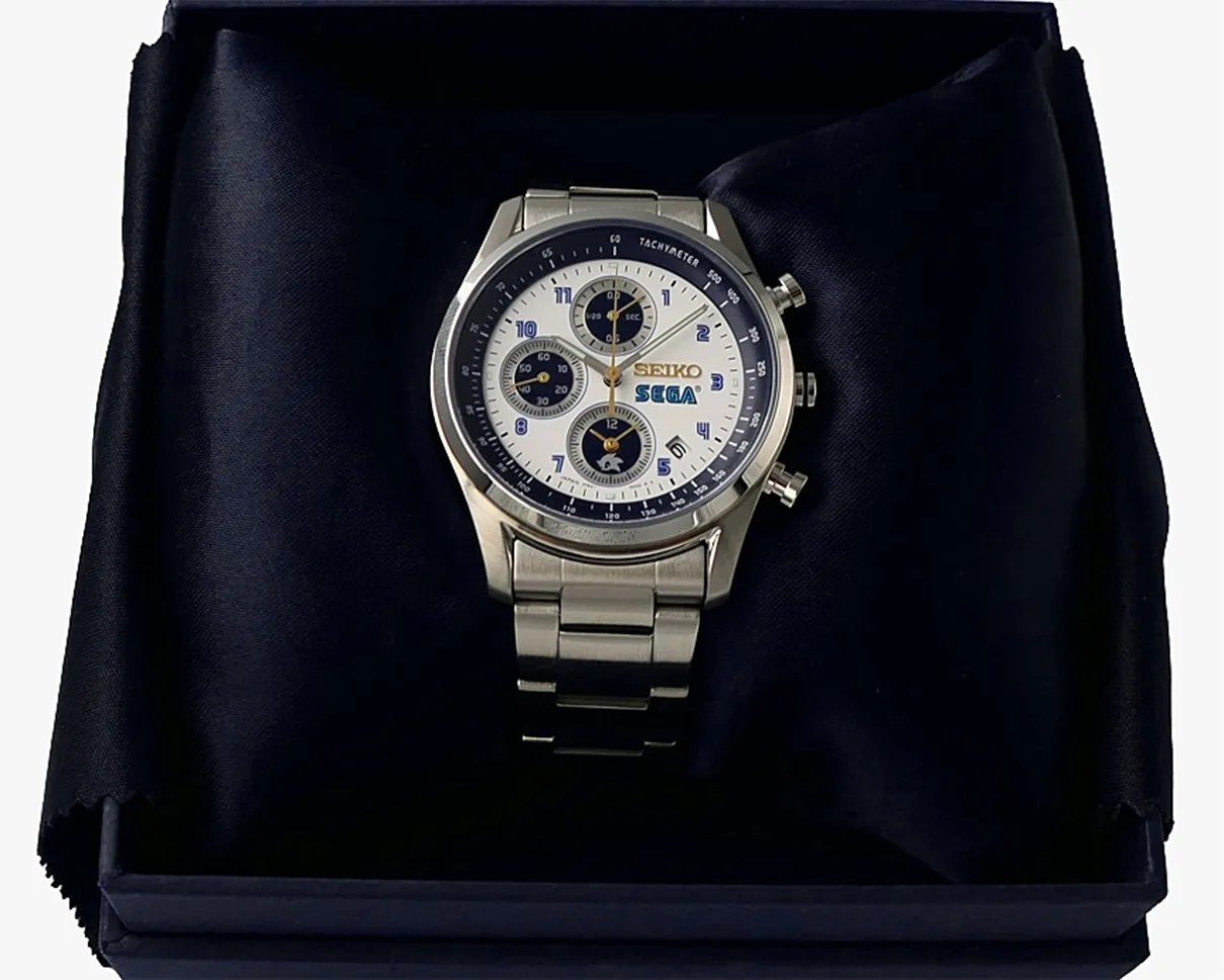 Silver Seiko wristwatch with white and blue dial featuring SEGA logo, displayed on black fabric inside a box.