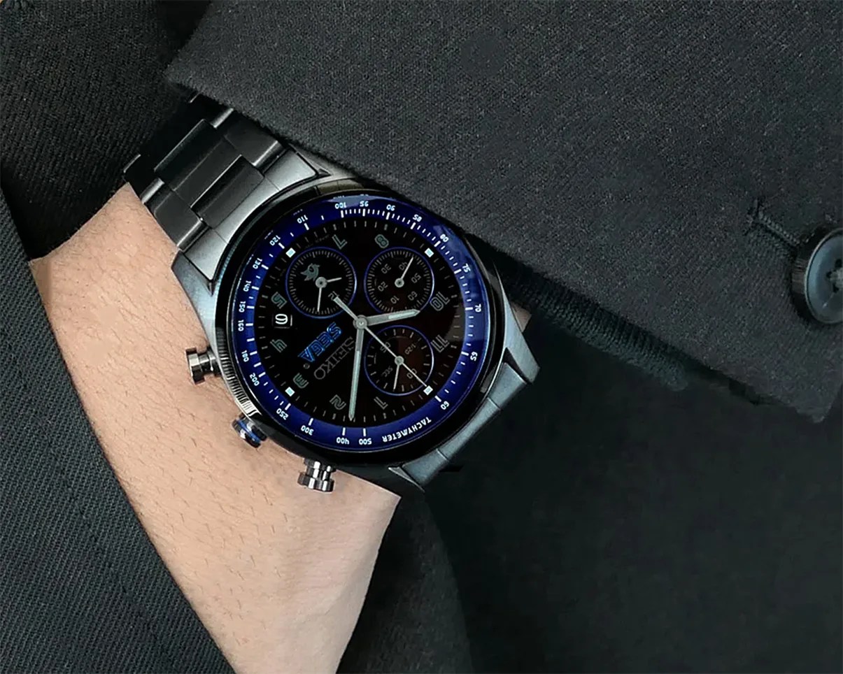 Black stainless steel chronograph watch with blue tachymeter bezel worn on a wrist under a black suit sleeve.