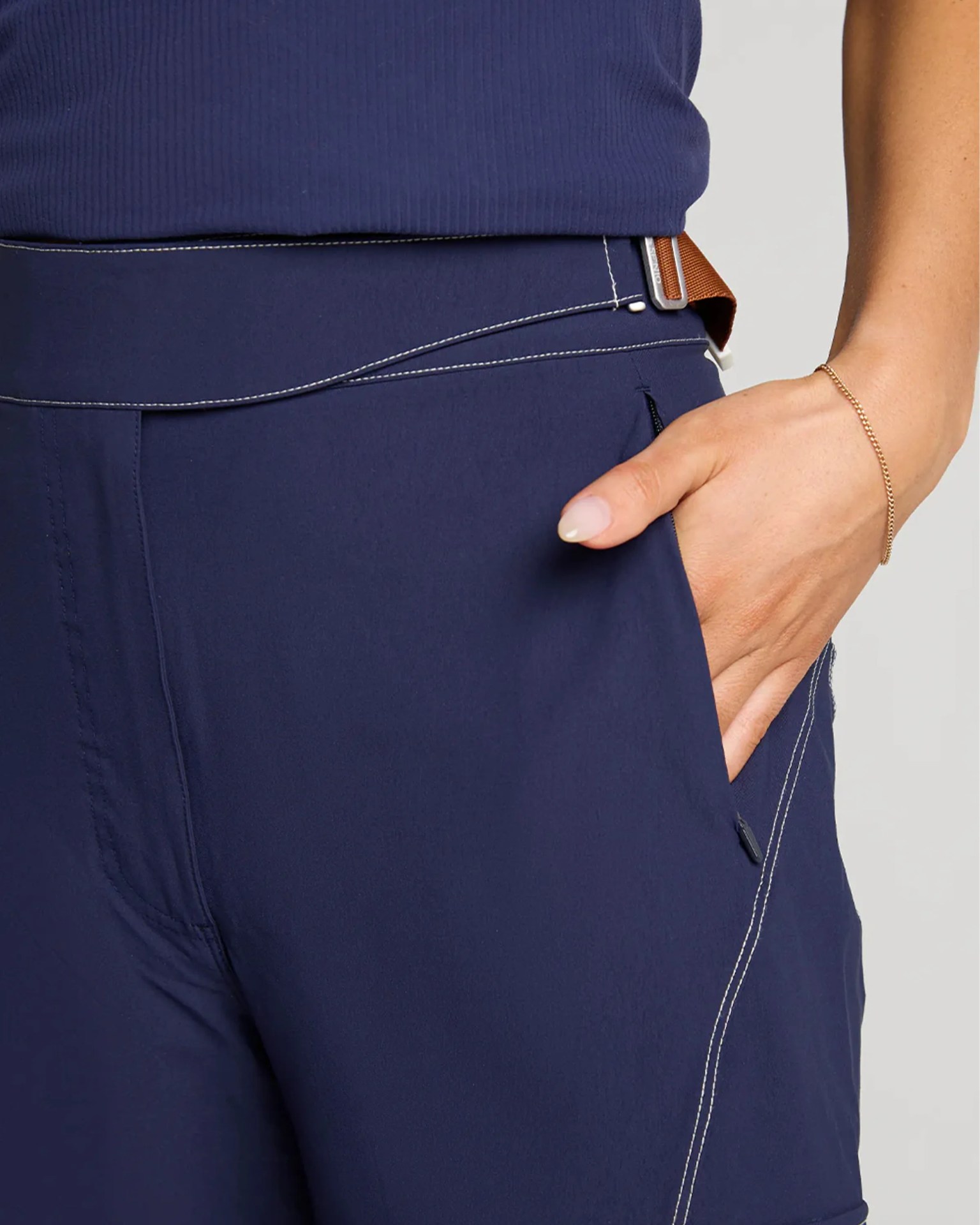Close-up of navy blue pants with white stitching and a hand in the pocket wearing a thin gold bracelet.