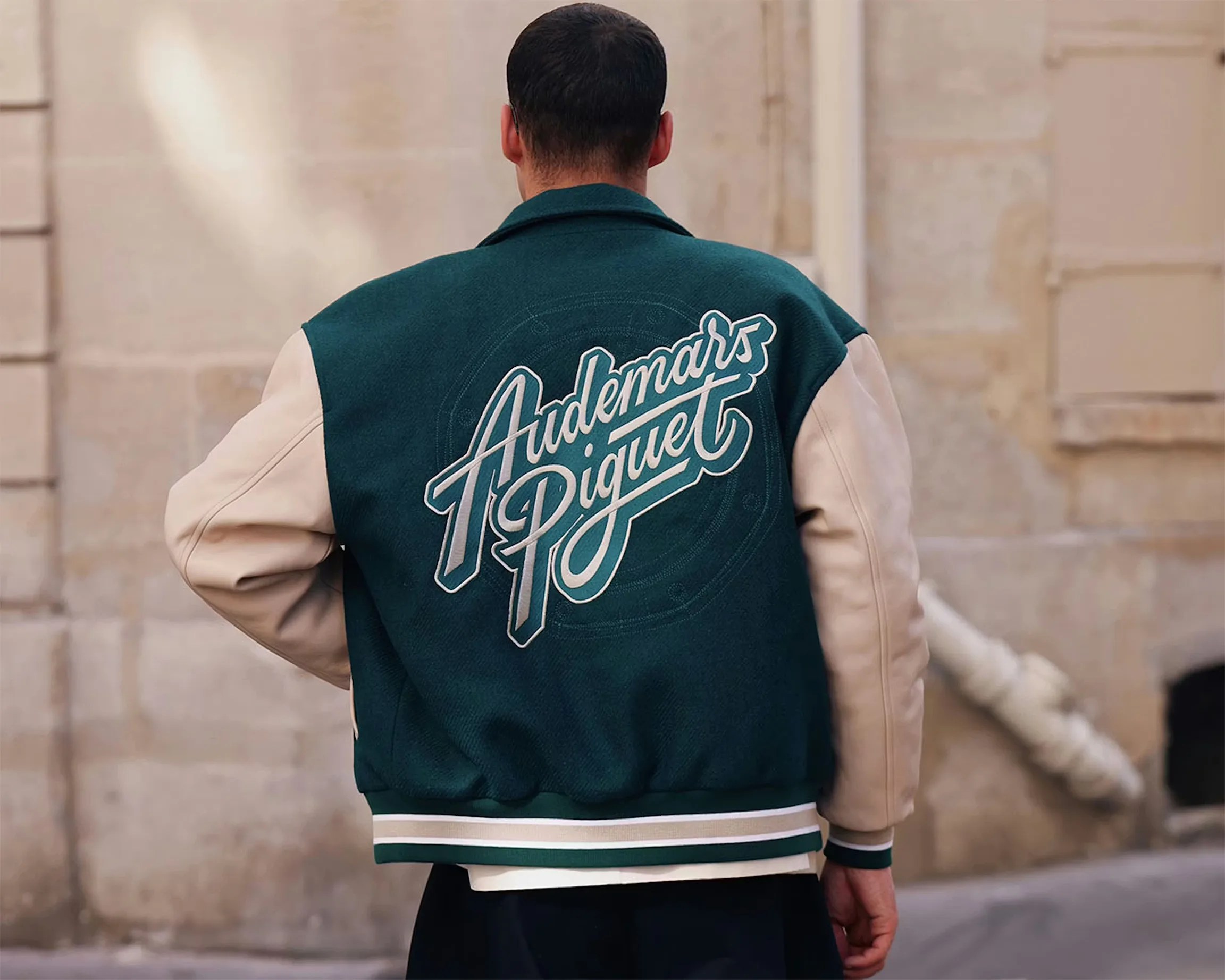 Man wearing a dark green and beige varsity jacket with "Audemars Piguet" embroidered on the back.