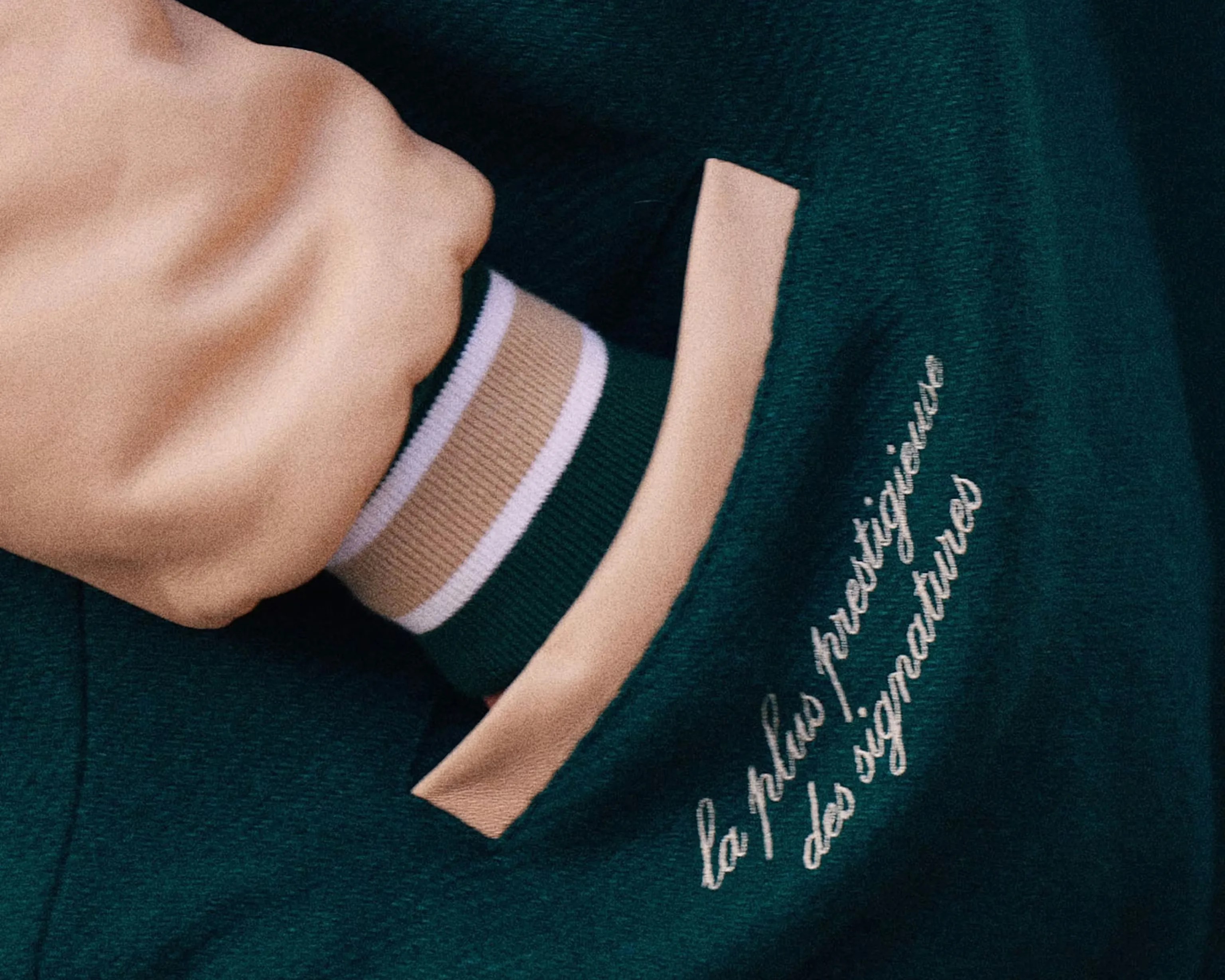 Hand in dark green jacket pocket with beige trim and embroidered French text "la plus prestigieuse des signatures.