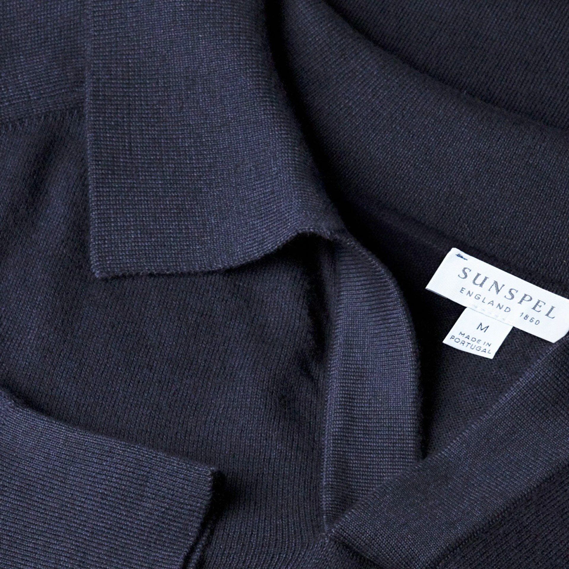 Close-up of a dark navy knit sweater with a folded collar and a visible Sunspel brand label.