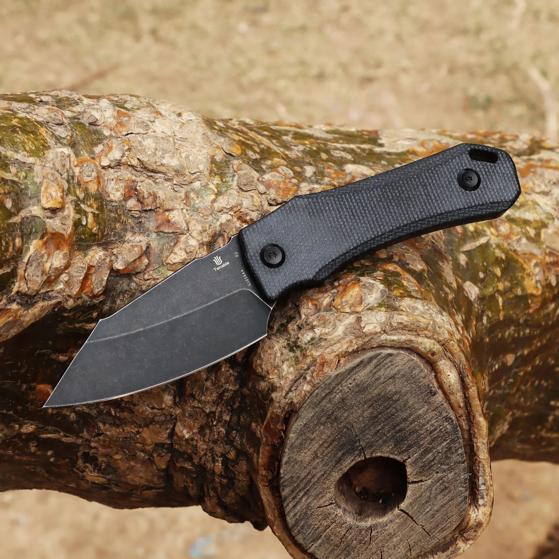 Black folding knife with textured handle resting on a tree branch.