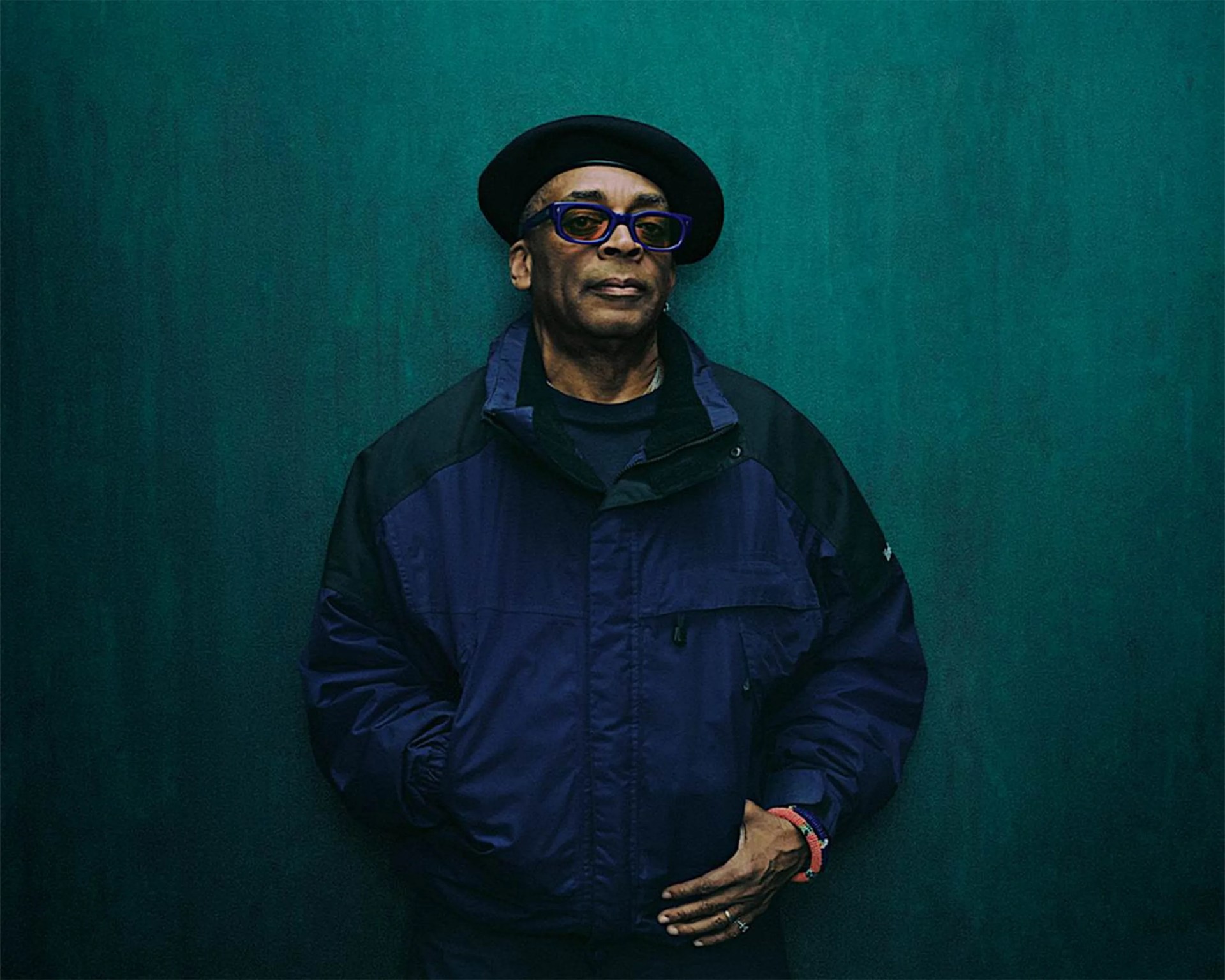 Man wearing a black beret, blue glasses, and a navy blue jacket against a dark teal background.