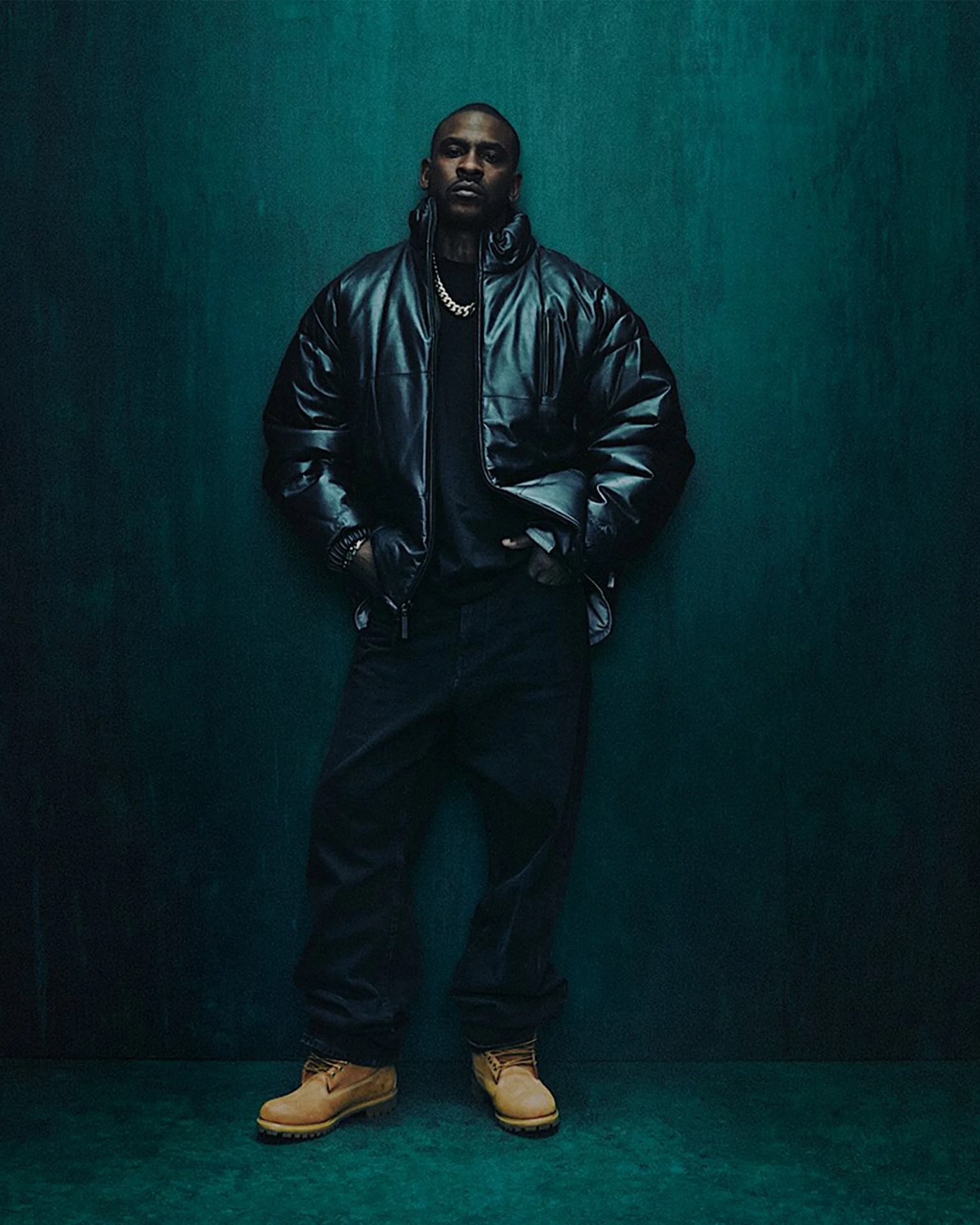 Man wearing a black leather puffer jacket, black pants, gold chain necklace, and tan work boots standing against a dark teal background.