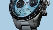 Close-up of a stainless steel chronograph watch with a light blue dial and black subdials on a light blue background.