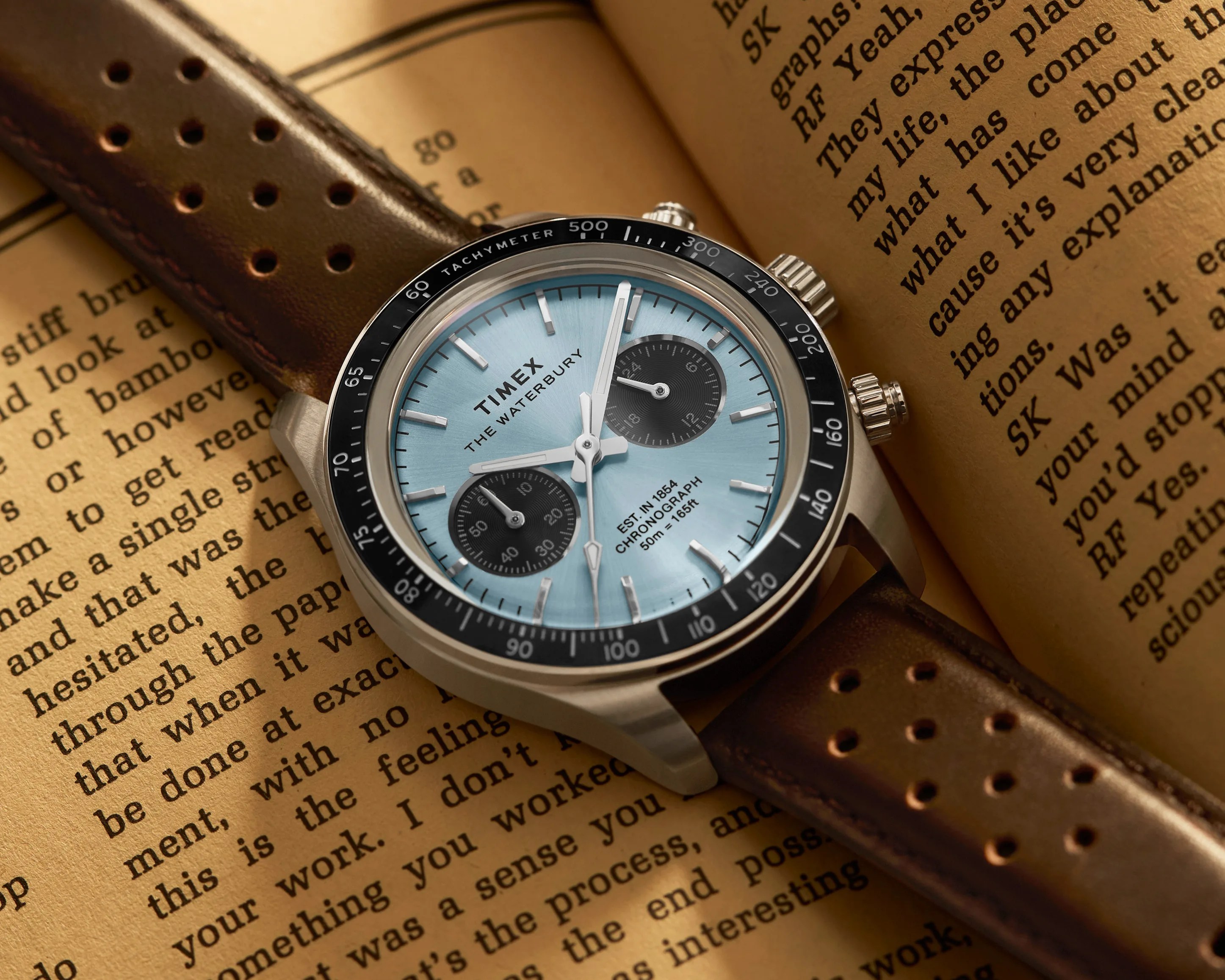 Timex Waterbury chronograph watch with light blue dial, black tachymeter bezel, and perforated brown leather strap on an open book.