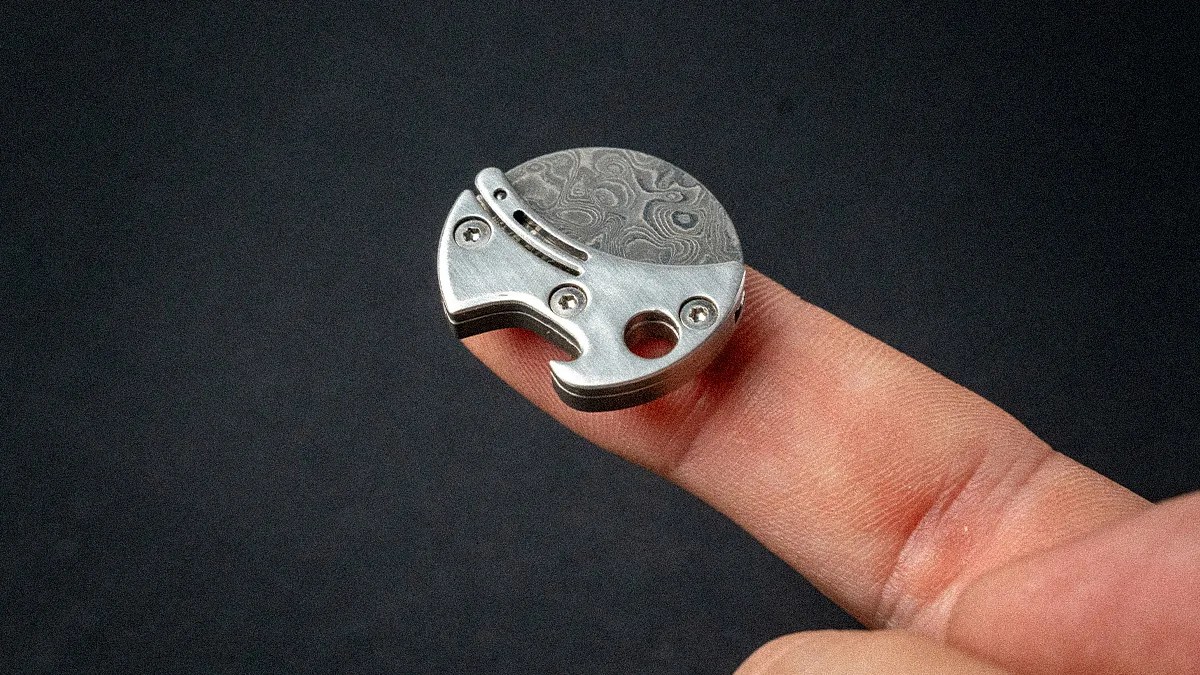 Small round metal object with screws and a patterned surface resting on a fingertip against a dark background.