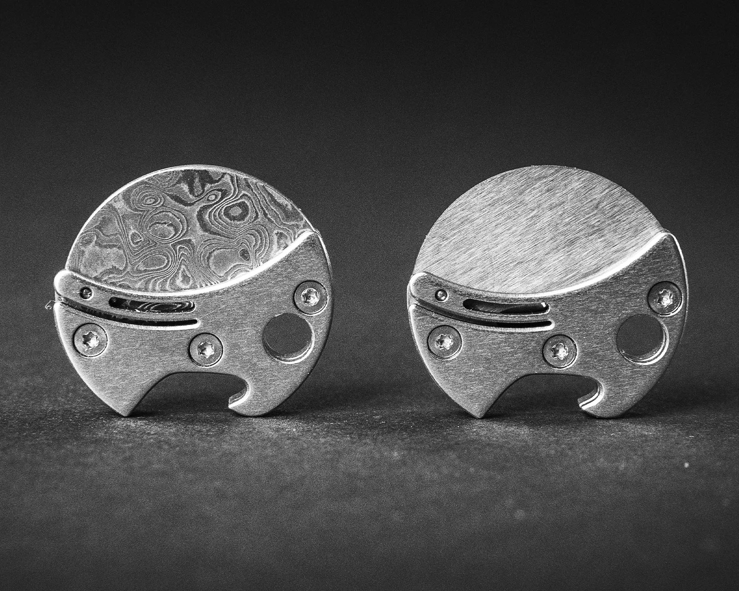 Two round metal objects with screws and a curved design, one with a patterned top and the other with a brushed finish, on a dark surface.