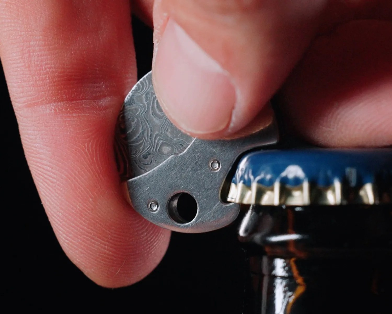 Hand holding a small patterned metal bottle opener removing a blue bottle cap from a dark glass bottle.