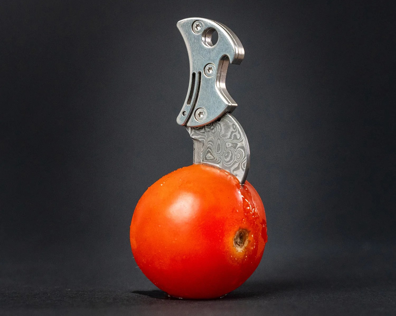 Small curved knife with a patterned blade stabbed into a red cherry tomato against a dark background.