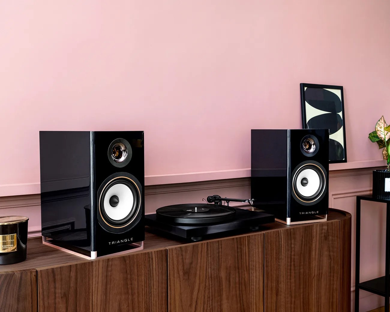 Pair of black Triangle bookshelf speakers with white cones on a wooden cabinet next to a black turntable.