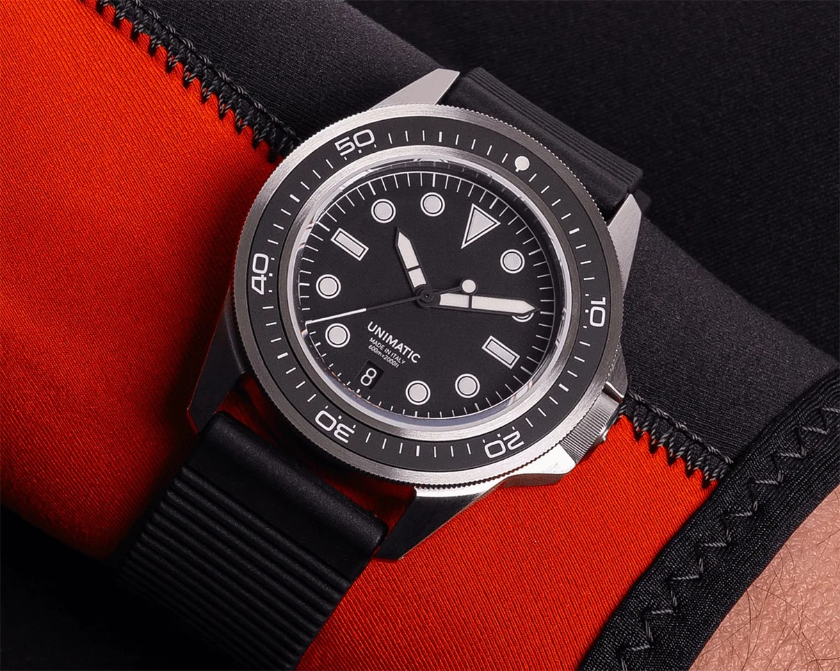 Black Unimatic dive watch with white markers and date window on a black rubber strap worn over a red and black wetsuit.