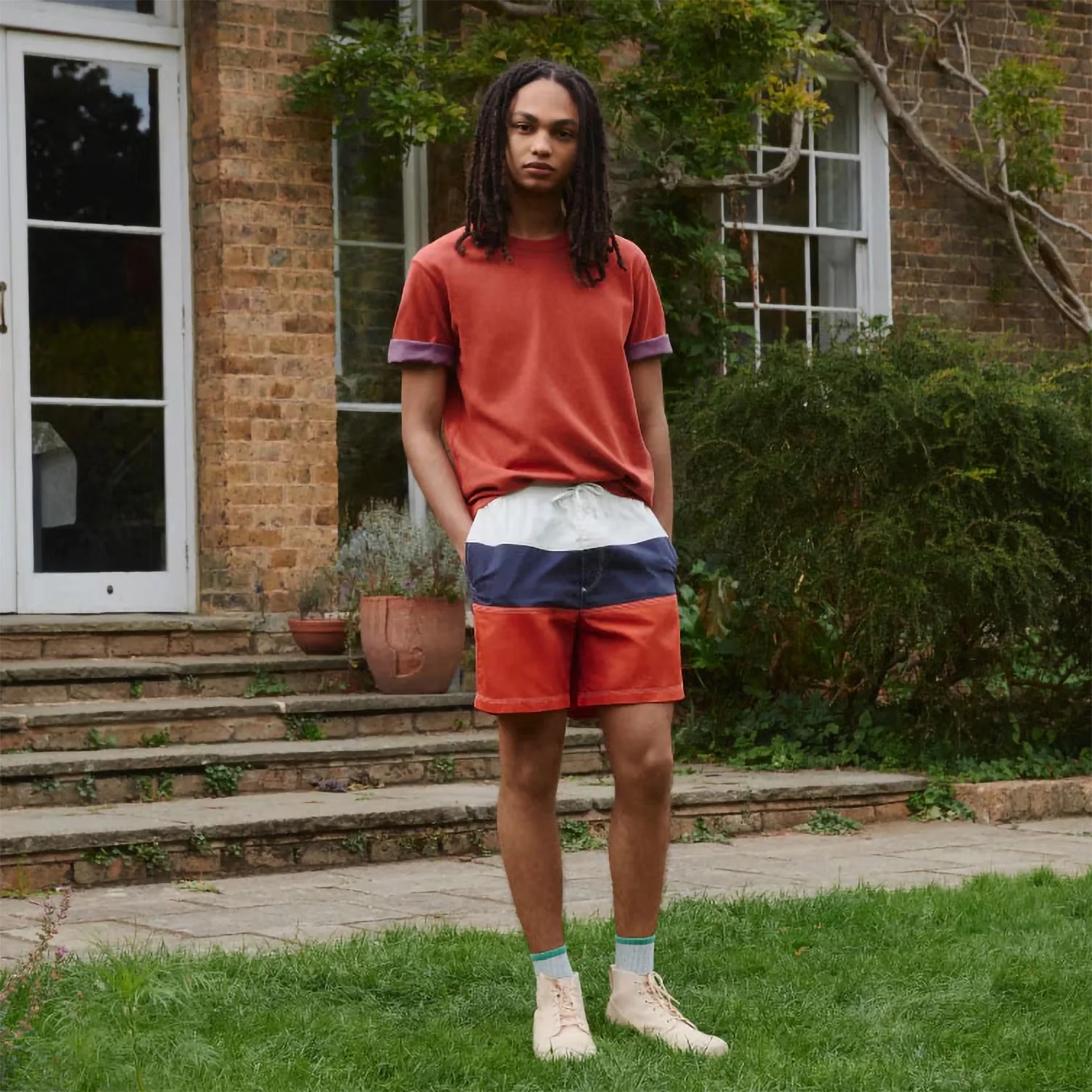 Person standing on grass wearing a rust-colored T-shirt with rolled sleeves, color-blocked shorts, and light beige high-top sneakers.