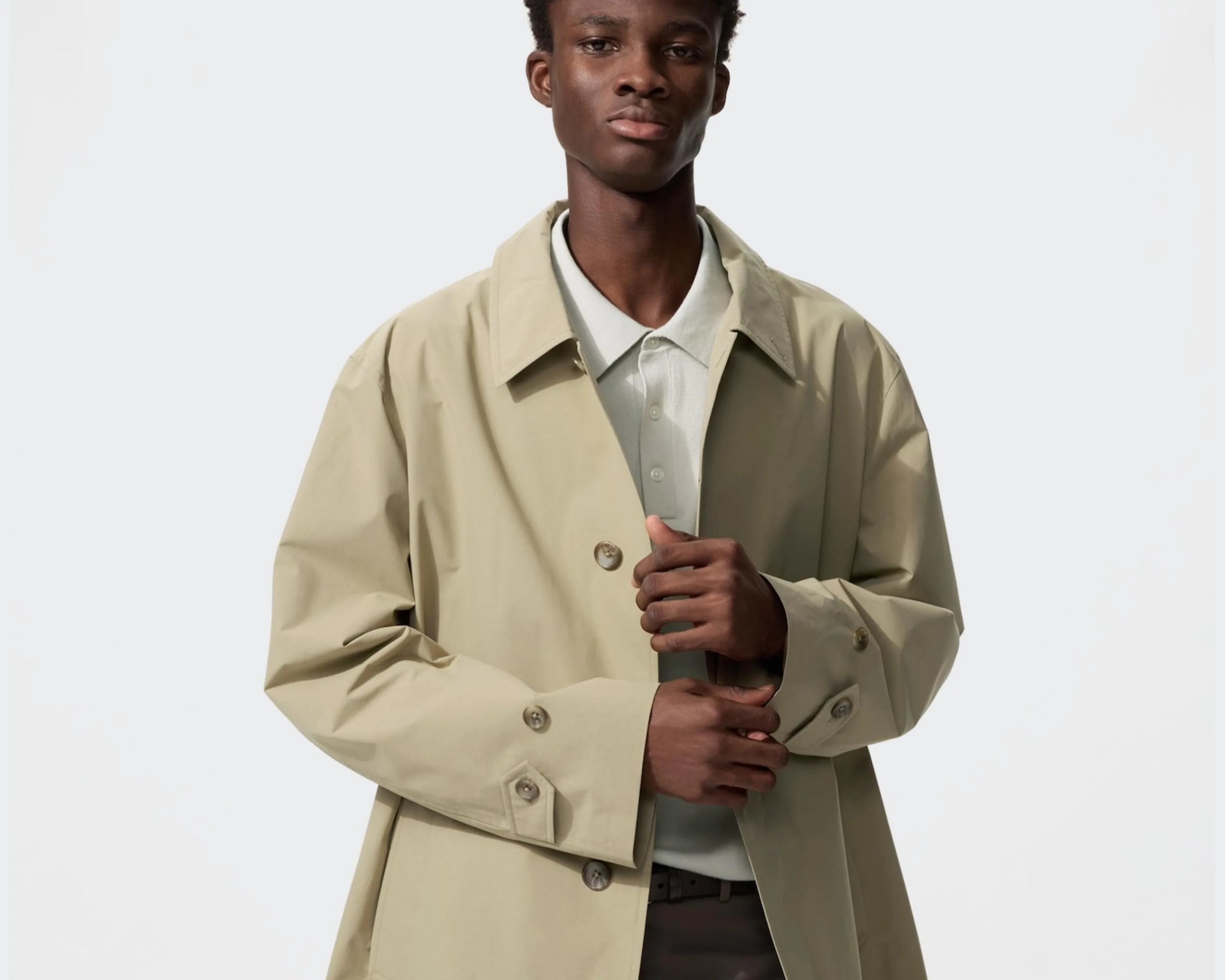 Man wearing a beige trench coat over a light gray collared shirt against a plain white background.