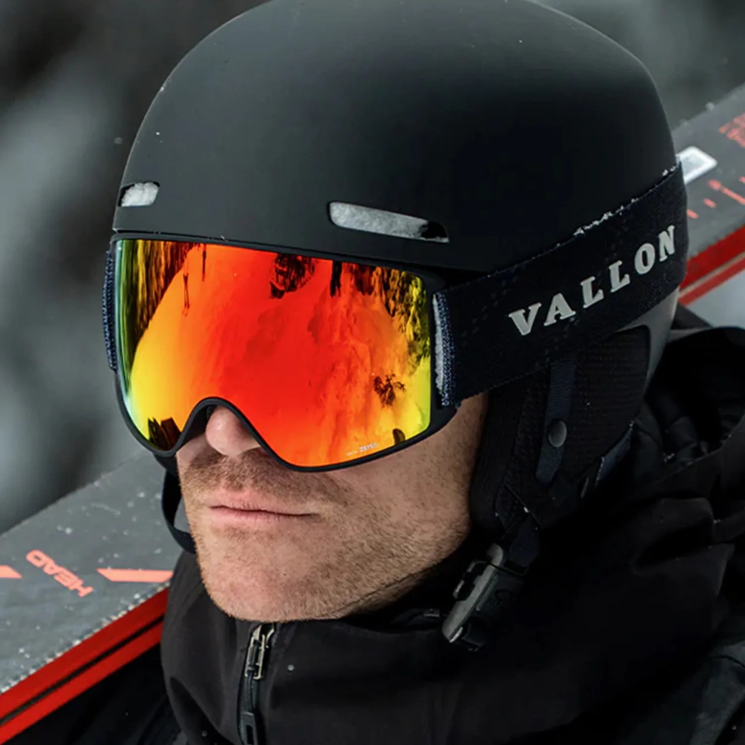 Man wearing a black Vallon ski helmet and orange reflective ski goggles with a ski in the background.